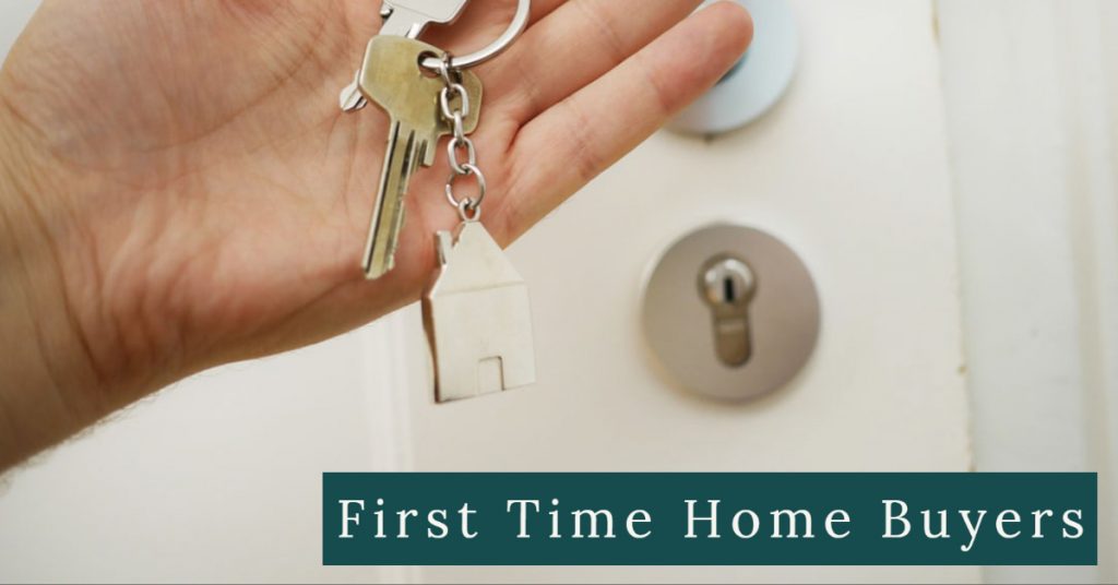 Image of a person's hand holding a set of house keys on a key chain in front of a door to a house. Captioned: First Time Home Buyers