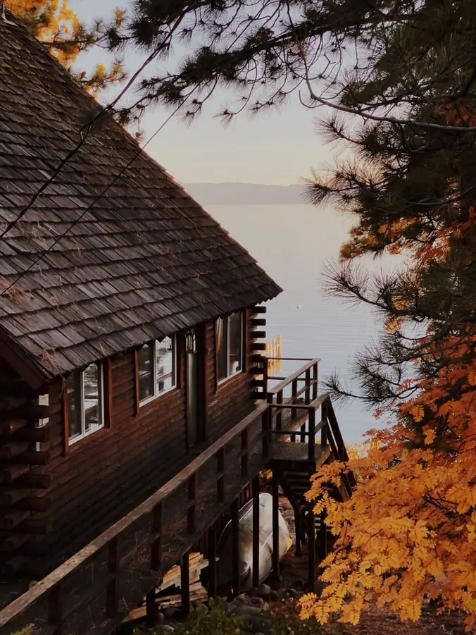 Image of a cabin style house surrounded by large trees with a lake and mountains in the background.