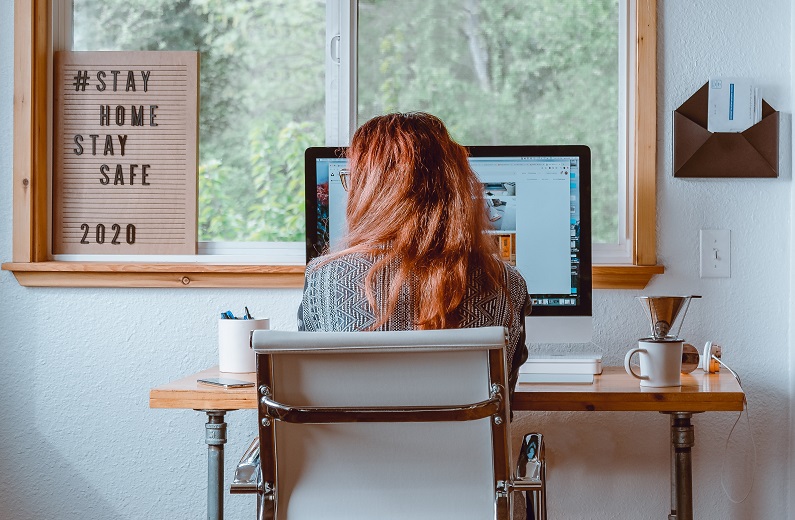Image of a person sitting in front of a computer on a desk looking out a window with a sign the reads stay home stay safe 2020