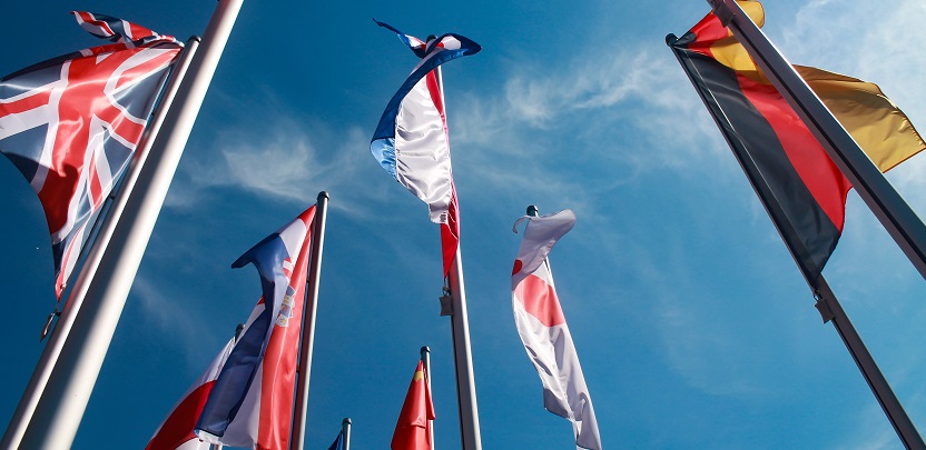 Image looking up at several international flags on flag poles with a blue sky in the background. Captioned: Immigration Status and Real Estate