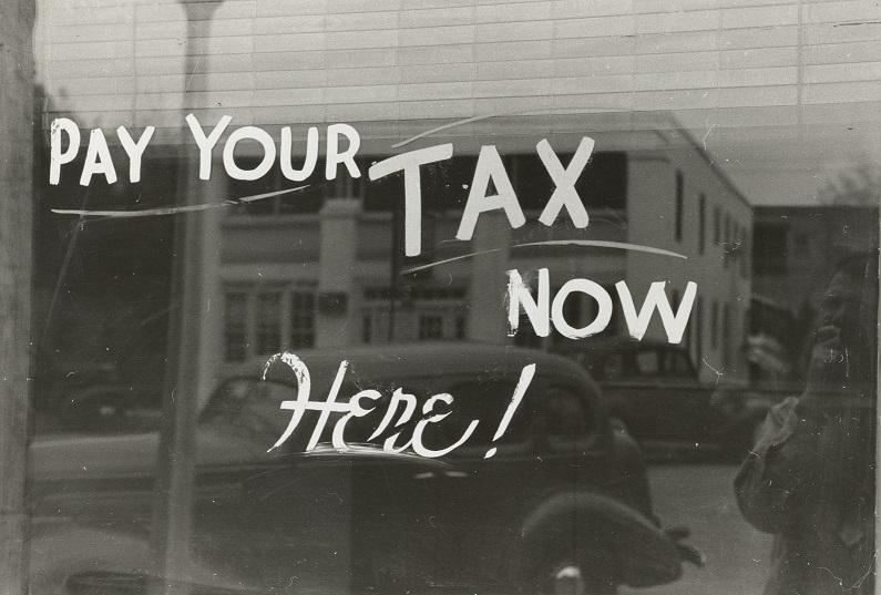 Historical image of a window on a downtown building with the words "Pay Your TAX Now Here!" painted on it. Captioned: Back Taxes Can Impact Your Real Estate Title