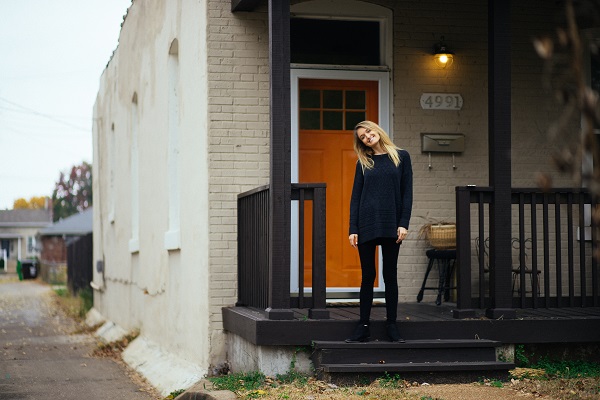 Image of a woman standing on the porch of a house in front of a door. Captioned: The Single Woman and Home Buying.