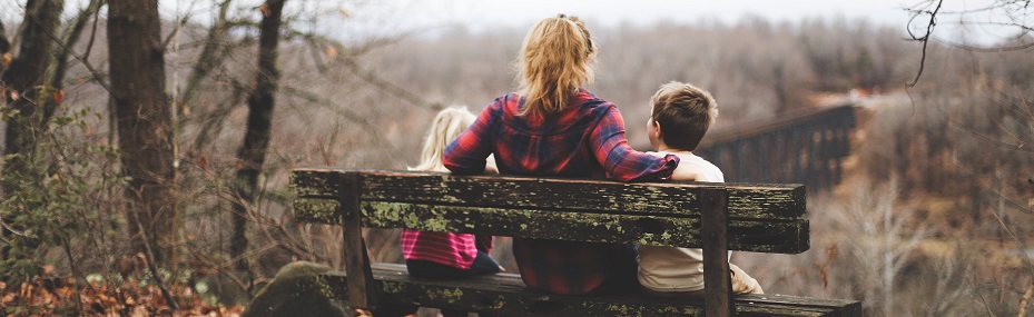 Image of a person sitting on a bench in a wooded area with two children. Captioned: Is It Time to Place Your Home in a Living Trust?