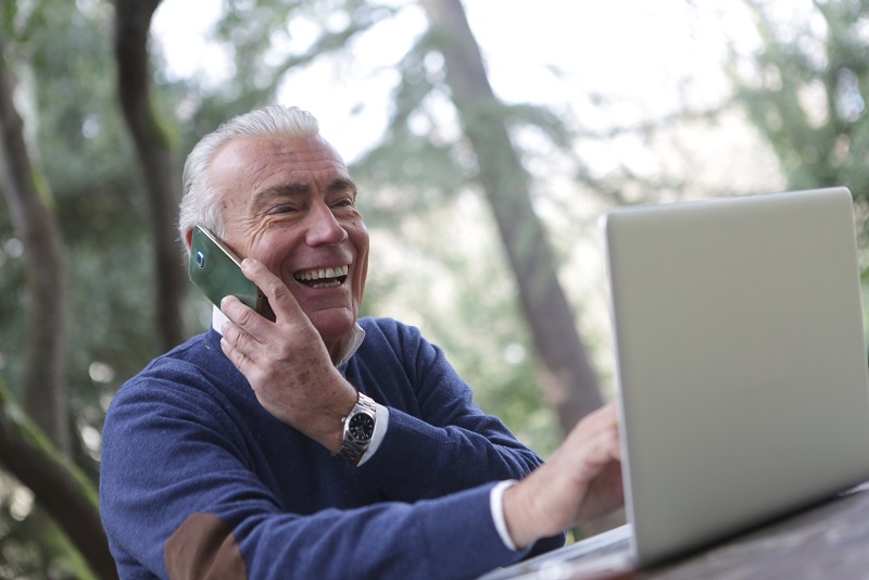 Image of a person sitting outside talking on a phone and using a computer at the same time. They seem very happy.