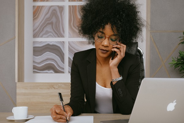 A person sitting at a table, talking on the phone while writing with a pen on a peice of paper.