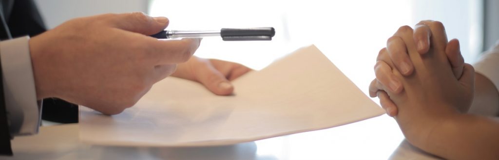 Image of legal documents being offered for signature from one person to another person seated at a table. Captioned: Selling Your Home? A Few Things to Know About Listing Agreements