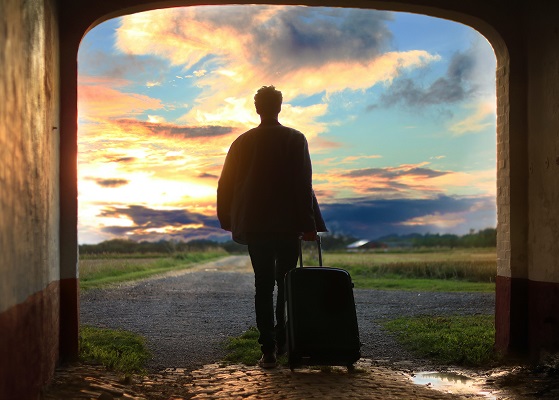 Image of a person with a suitcase standing in a doorway gazing into large outdoor scene with blue sky at sunset.