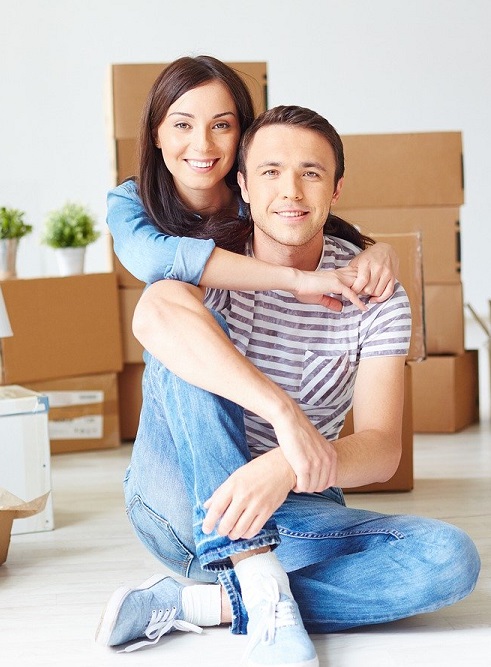 Two people sitting in a house with moving boxes behind them,