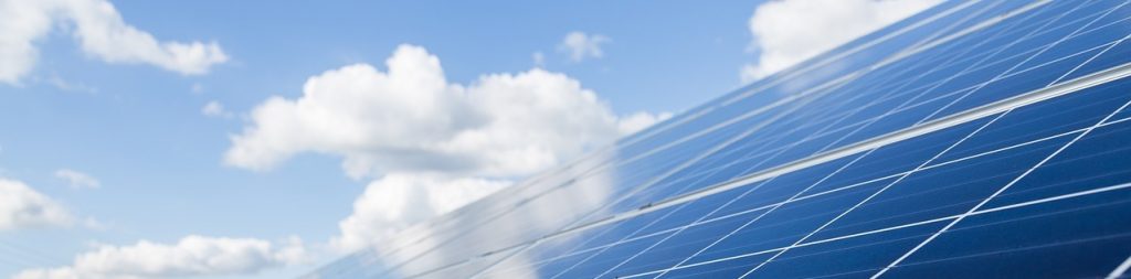 Image of solar panels facing a bright blue sky with some fluffy white clouds floating by.
