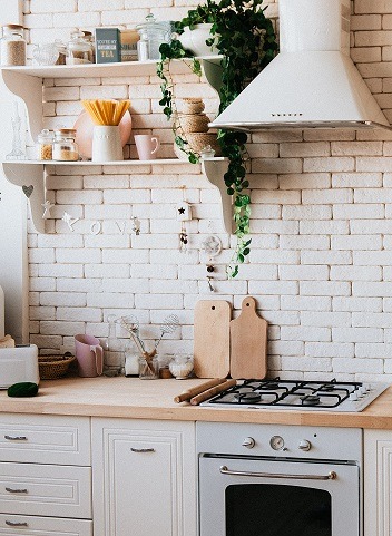 Close image of the kitchen area of a house showing a stove, oven, counter, and some cabinets and shelves. A nice exposed brick wall in the background.