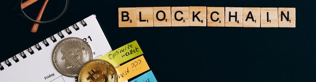Image of letter tiles that spell out blockchain on a black surface with some reading glasses, a part of a paper calendar, and some coins made to look like bitcoins.
