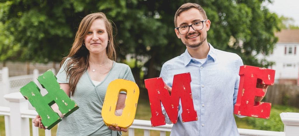 Two people standing outside holding letters to form the word HOME.
