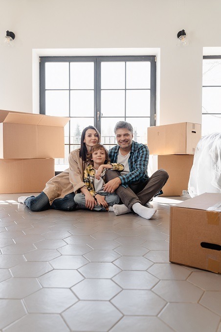 Image of a family sitting on the floor of a house with moving boxes around them, seemingly happy to be moving into a new home.