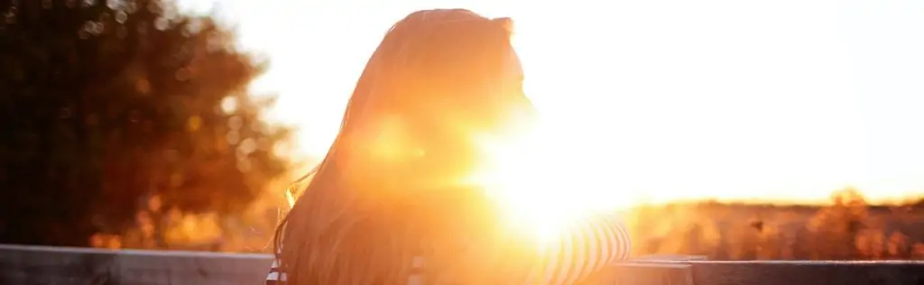 A person in deep thought, leaning on a fence with a bright sunset backlighting them.
