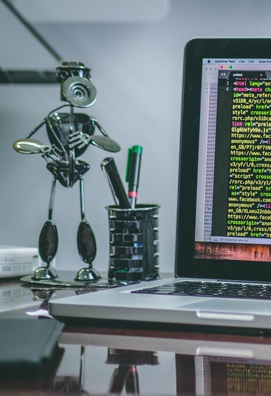 Software robots are like people. Image of a steampunk type robot sculpture sitting on a desk next to a computer with programing code on the screen.