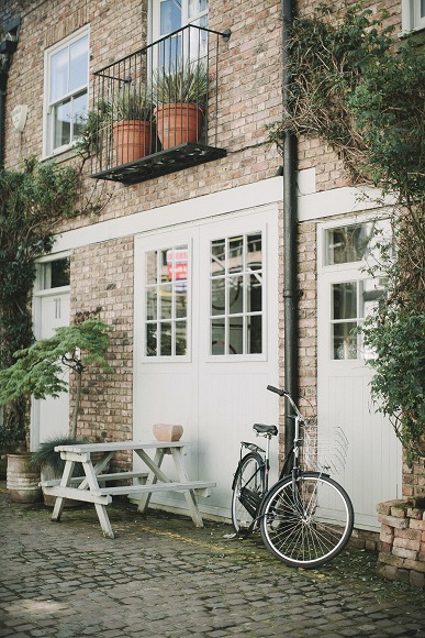 Outside of a residential building, natural brick exposed with large white doors. picnic table and bicycle sitting on a cobblestone area