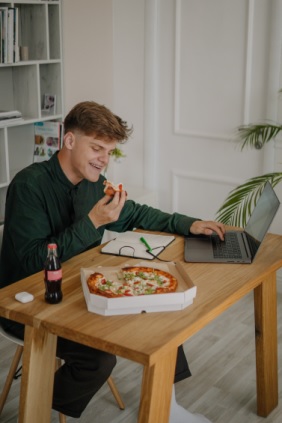 A young man in a casual green shirt enjoys a slice of pizza while working on his laptop at a wooden desk in a cozy home setting. The scene highlights the comfort and convenience of working from home, juxtaposing the ease of indulging in personal comforts while managing work tasks.